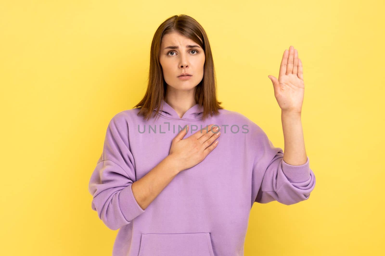 Serious conscious woman holding one hand on chest and raising another swearing, trust and honest, patriotism, wearing purple hoodie. Indoor studio shot isolated on yellow background.