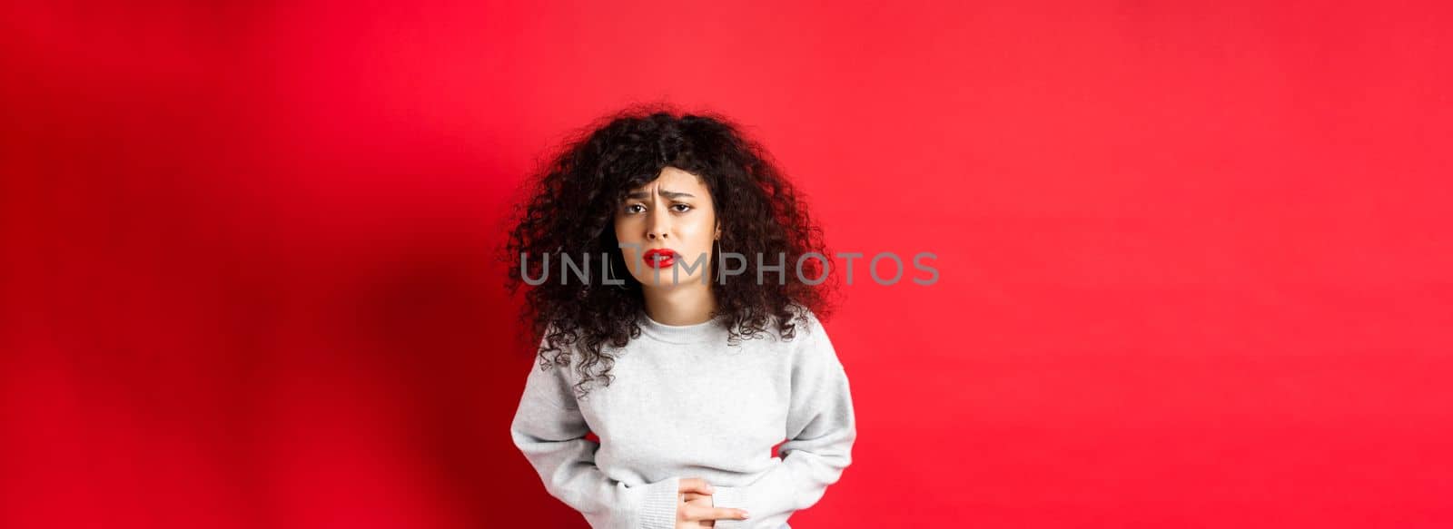 Image of young woman having stomach ache, bending from pain and complaining on painful menstrual cramps, standing on red background.