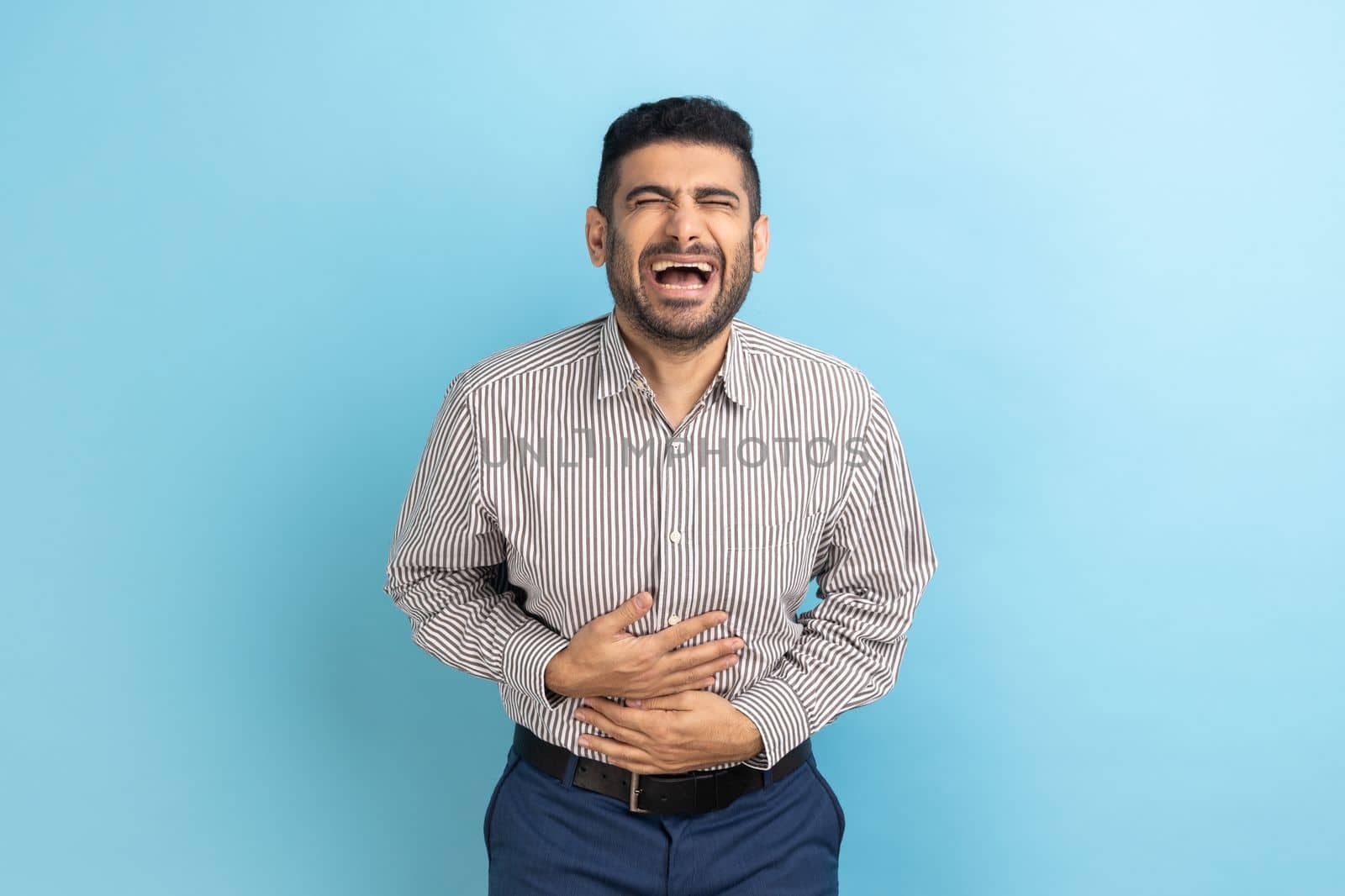 Portrait of funny handsome bearded young businessman standing with closed eyes and laughing, holding his belly, wearing striped shirt. Indoor studio shot isolated on blue background.