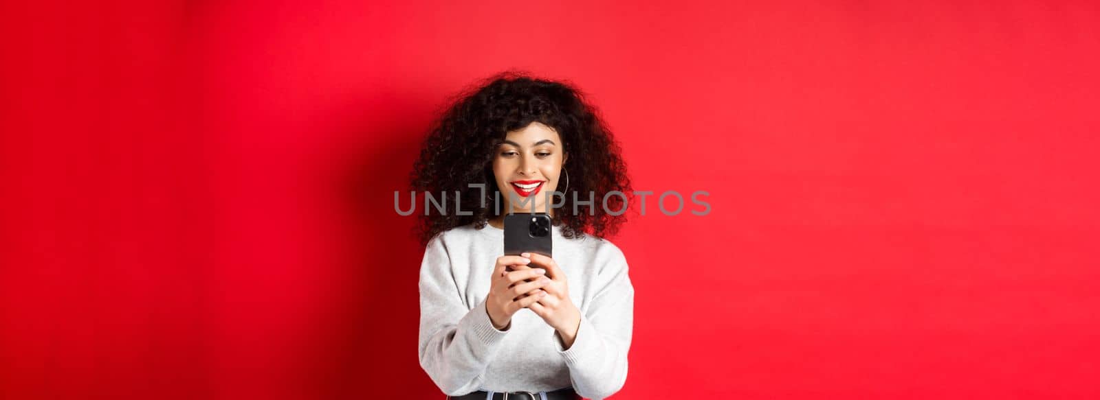 Smiling modern girl taking photos on smartphone, looking at screen and recording video, standing against red background.