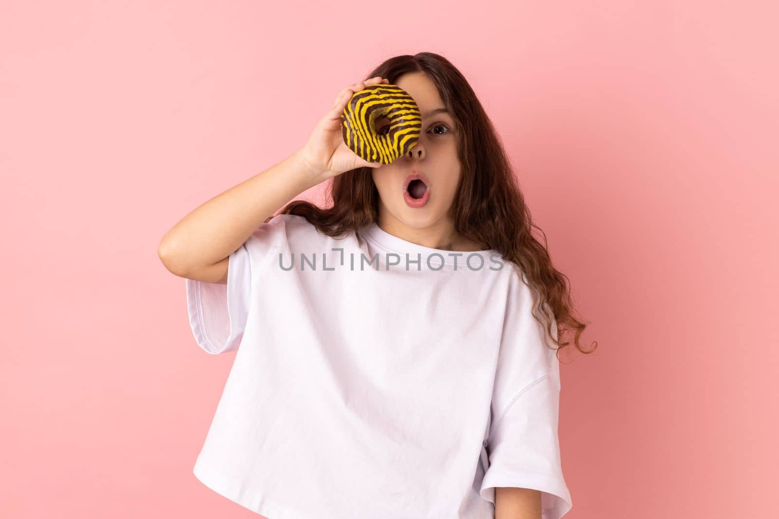 Portrait of excited amazed little girl wearing white T-shirt standing having fun, holding and covering eye with donut and keeps mouth open. Indoor studio shot isolated on pink background.