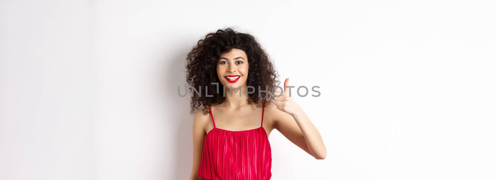 Cheerful woman with red lips and elegant dress, showing thumb up and smiling, recommending product, standing over white background.