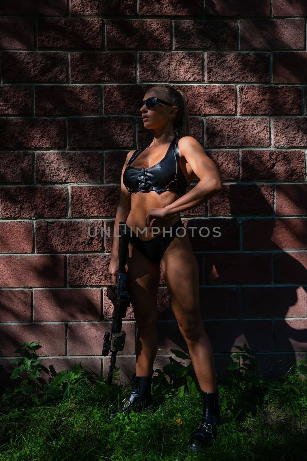 A caucasian woman in a bikini holds a machine gun against a brick wall