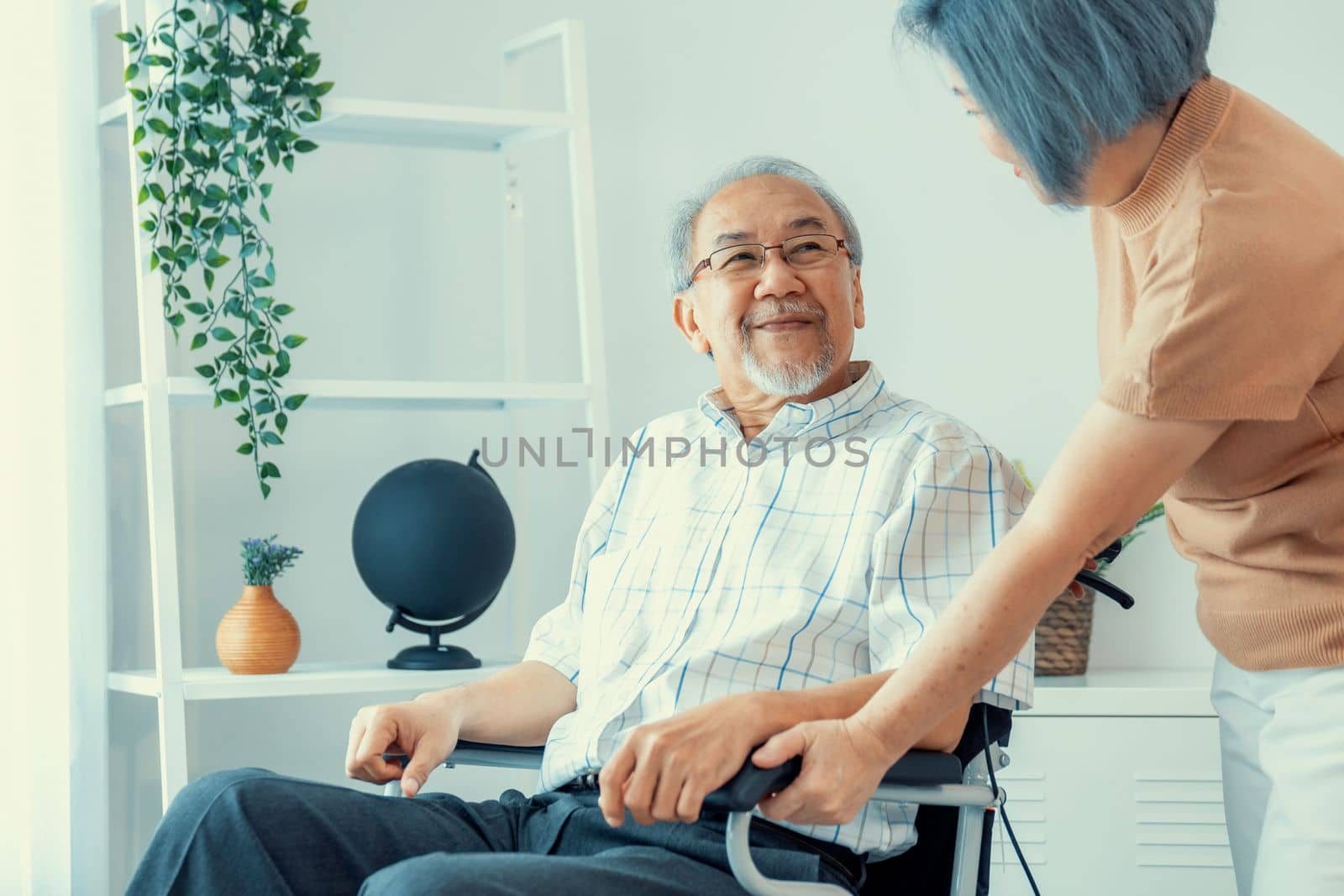 Senior wife giving support to her husband in his wheelchair with love, contented pensioner life. A senior couple is understanding and smiling at each other.