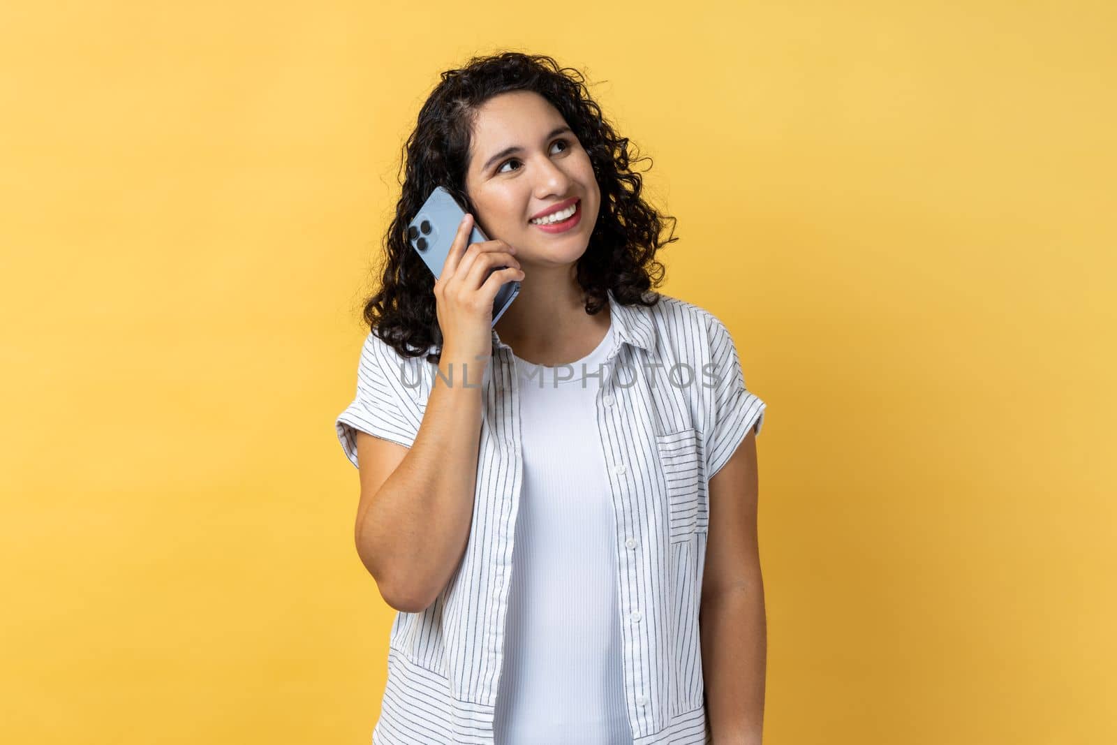 Portrait of smiling satisfied young adult beautiful woman with dark wavy hair standing and talking on smartphone, hearing good news. Indoor studio shot isolated on yellow background.
