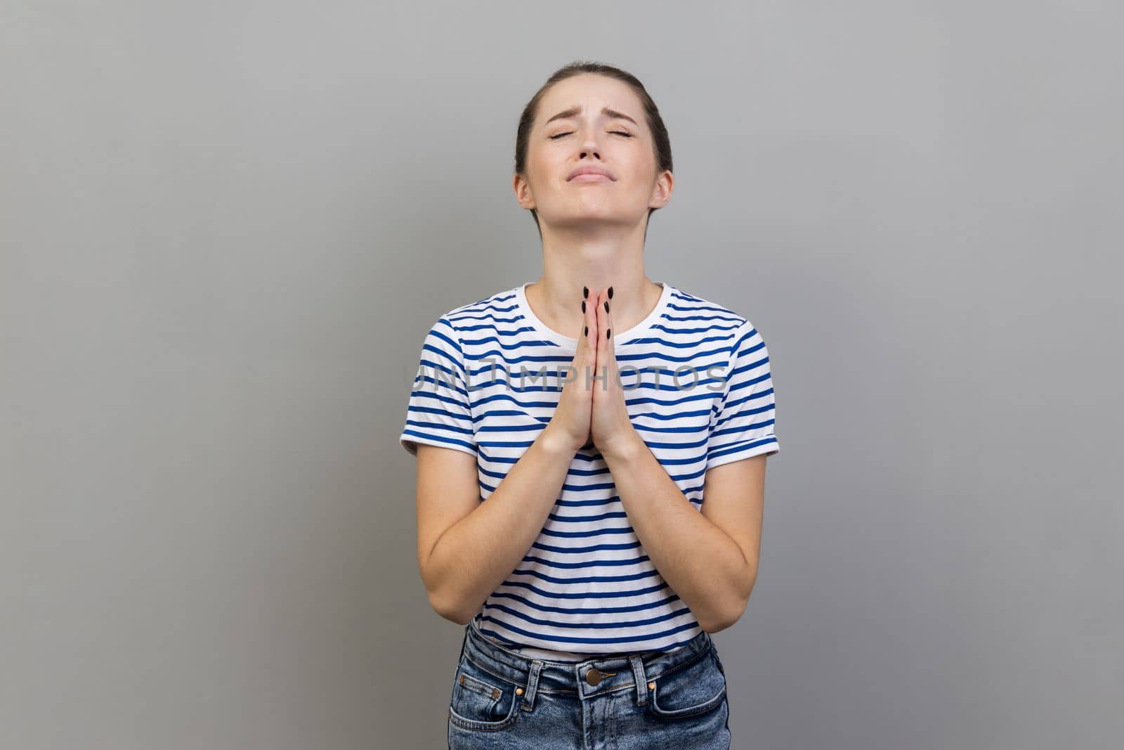 Please, I'm begging. Portrait of upset worried woman wearing striped T-shirt looking up with imploring desperate grimace, praying to god asking for help. Indoor studio shot isolated on gray background