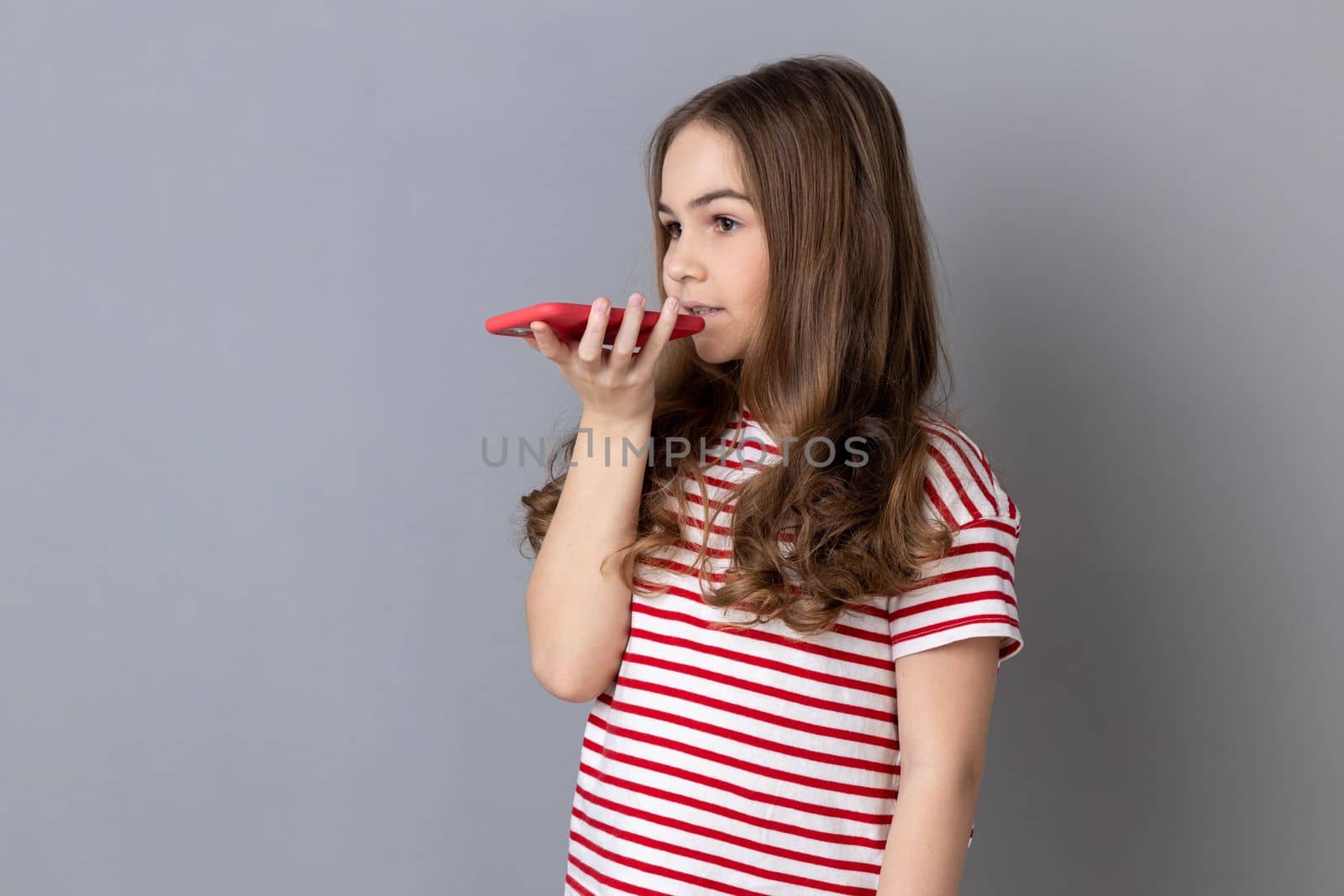 Virtual assistant. Little girl in striped T-shirt talking to smartphone using digital voice app, recording message, searching information on internet. Indoor studio shot isolated on gray background.