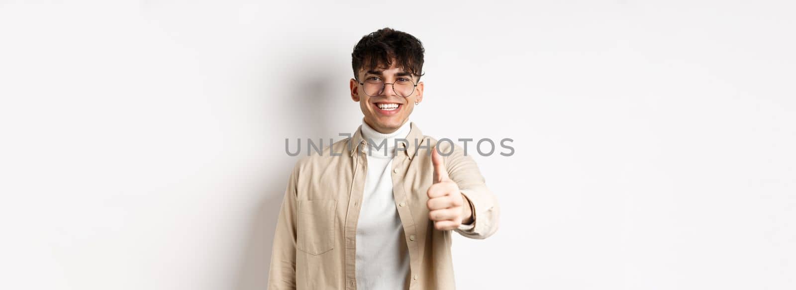 Image of happy young man showing thumb up and smiling satisfied, praise good job, say well done and look pleased, standing on white background.