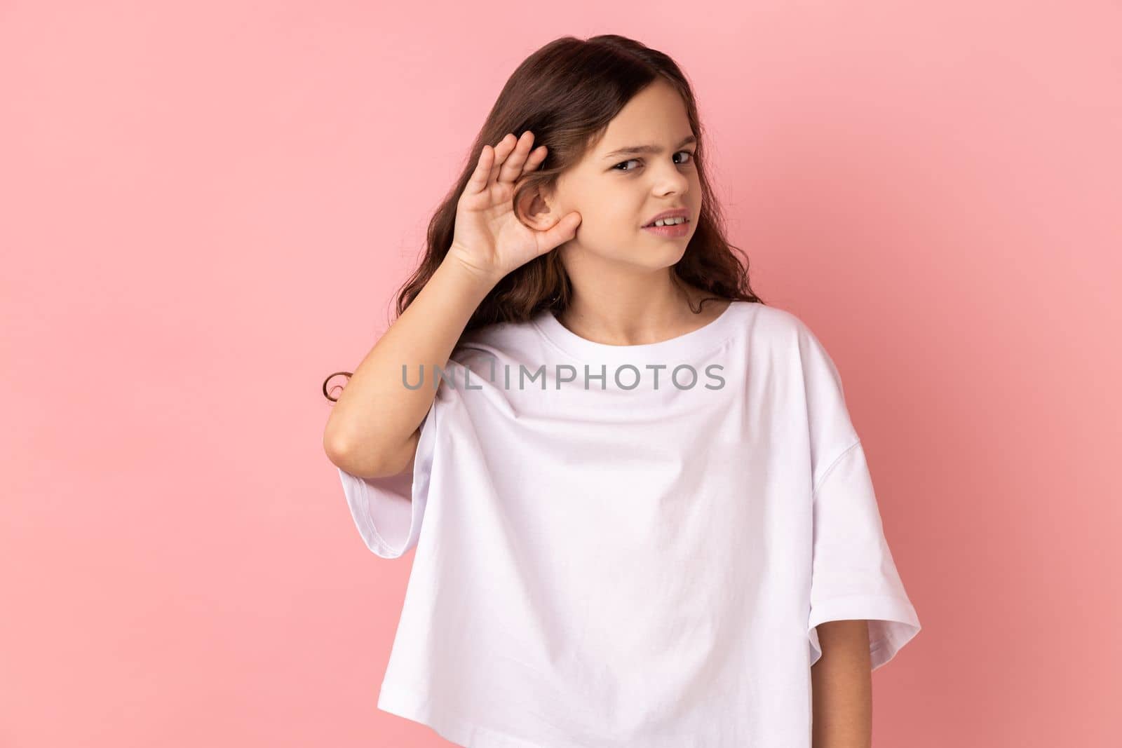 Portrait of curious little girl wearing white T-shirt holding hand near ear trying to hear secrets and find out gossips, listen to private whispers. Indoor studio shot isolated on pink background.