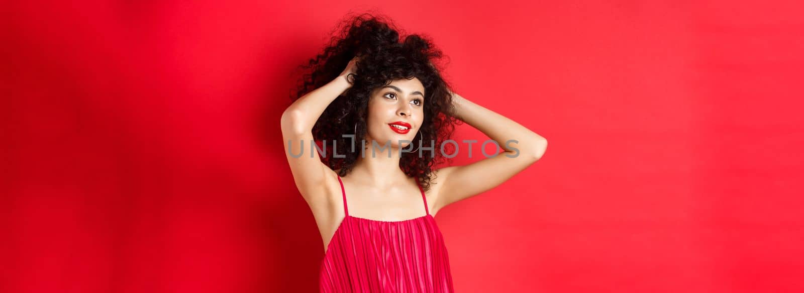 Carefree female model playing with hair, looking dreamy and relaxed at logo, smiling sensual, standing on red background.