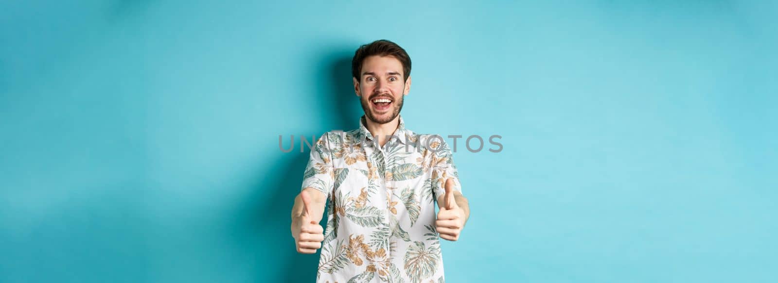 Excited smiling guy in hawaiian shirt show thumbs up, looking amazed, checking out cool promo, standing on blue background.