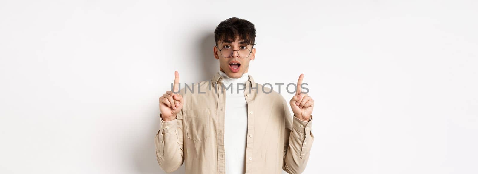 Amazed and excited man in glasses showing awesome promo deal, pointing fingers up and open mouth in awe, standing against white background.