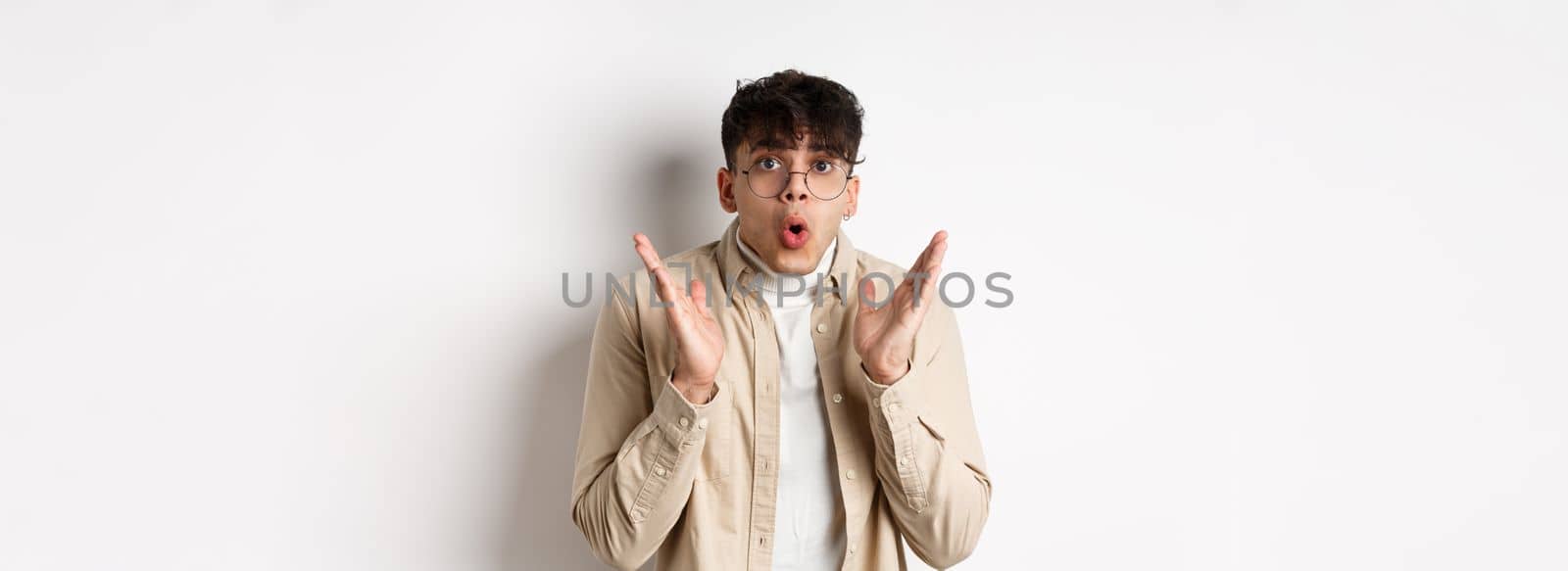 Portrait of surprised hipster guy in glasses checking out awesome promo, say wow and gasping amazed, raising hands up with disbelief, standing on white background.