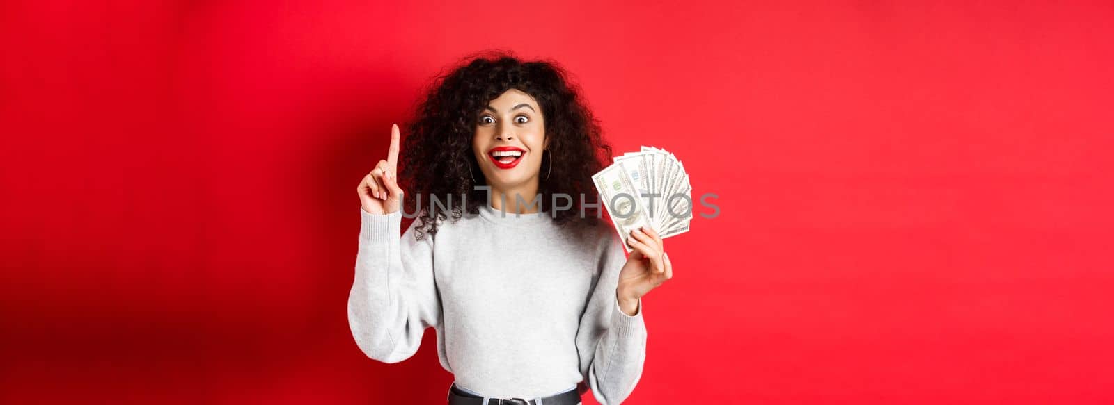 Young excited woman having an idea how make money, showing dollar bills and raising finger in eureka sign, standing on red background.
