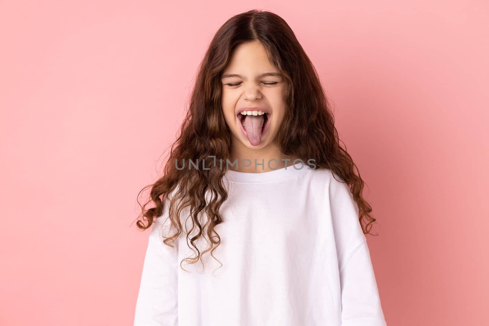 Portrait of childish carefree little girl wearing white T-shirt showing out tongue and closing eyes with naughty disobedient grimace, making face. Indoor studio shot isolated on pink background.