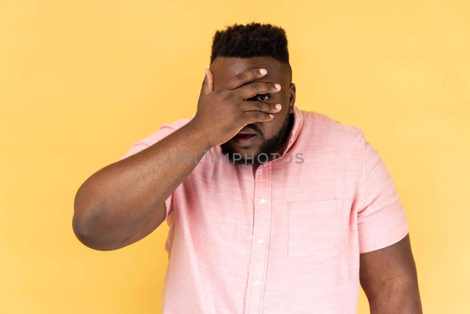 Portrait of handsome man wearing pink shirt looking through fingers, peeking with calm face, having suspicious, watching secrets. Indoor studio shot isolated on yellow background.