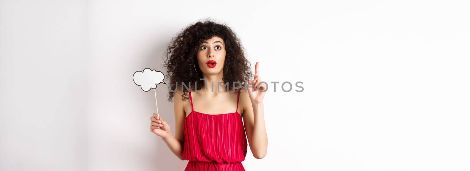 Excited fashionable woman in red dress pitching an idea, raising finger in eureka sign, holding thought cloud, standing on white background.
