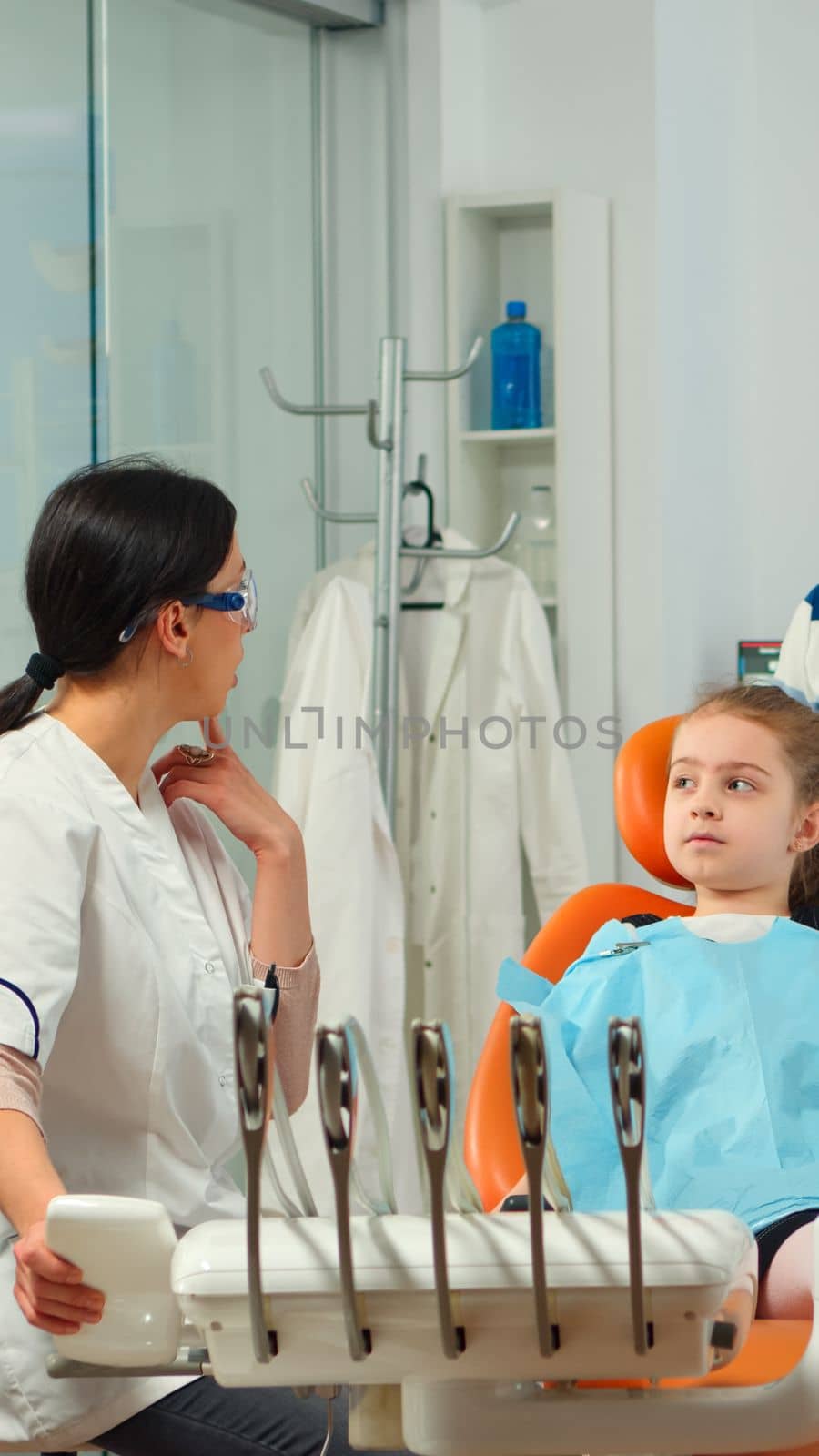 Stomatologist doctor explaining to mother the dental process of intervention for teeth problems of kid, girl showing to affected mass. Orthodontist speaking to child sitting on stomatological chair