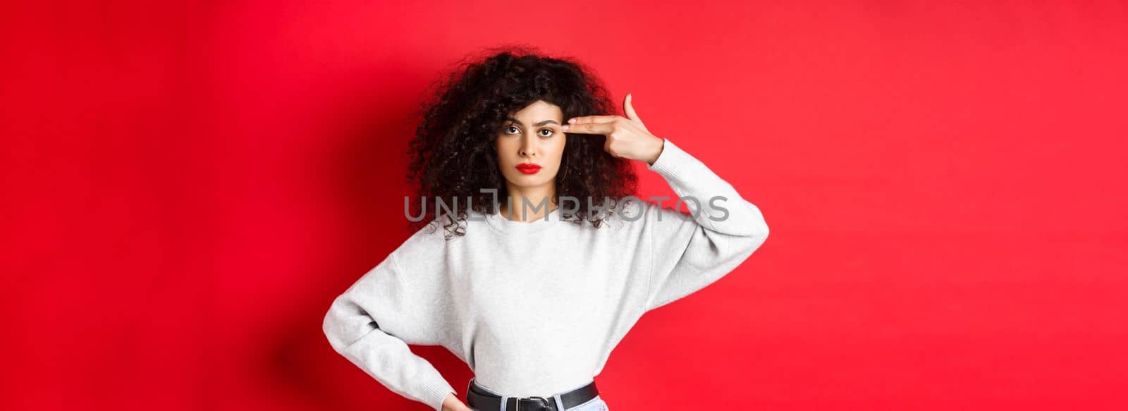 Annoyed caucasian woman with curly hair, showing hand gun sign on head, blow her mind off, standing bothered against red background.
