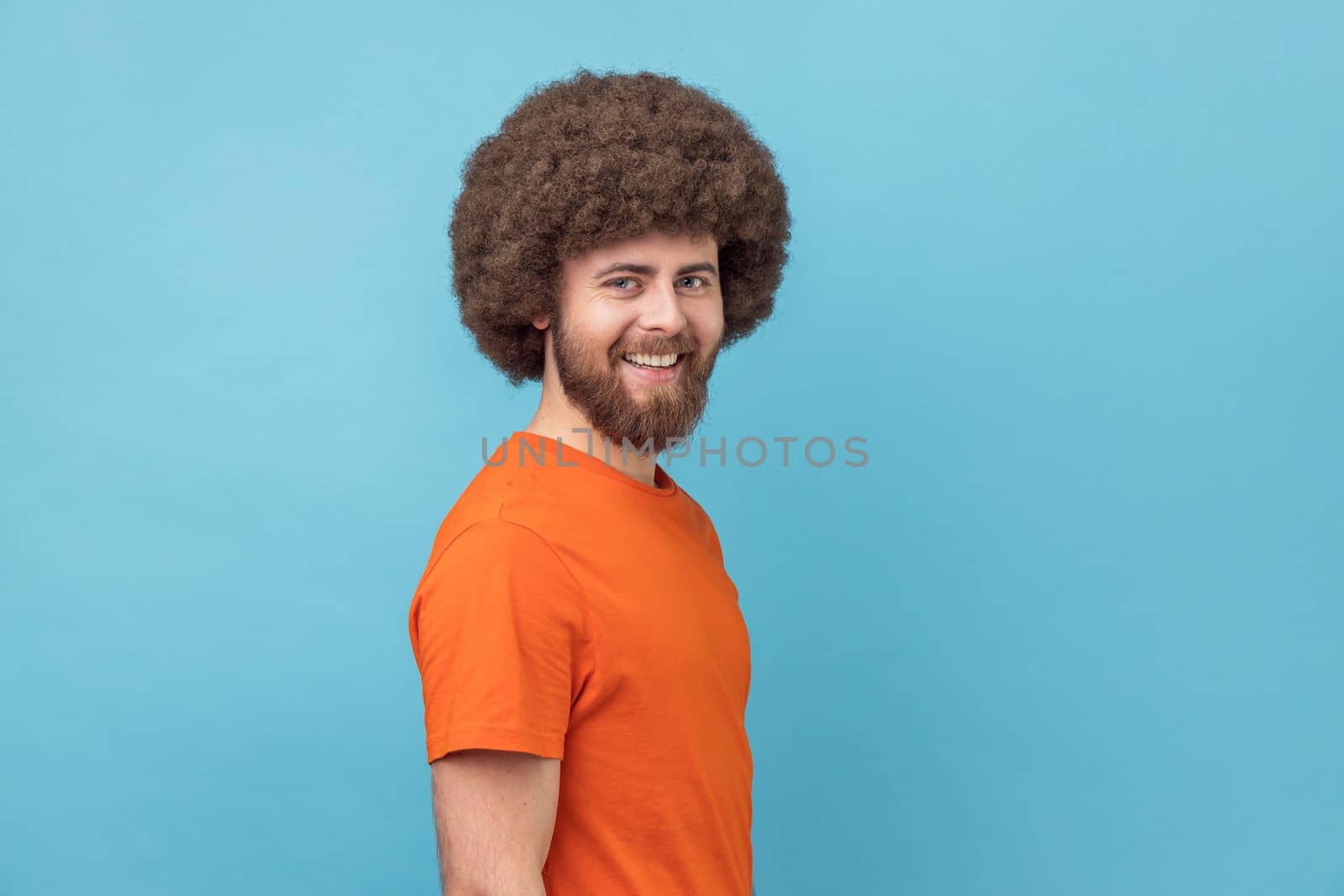 Side view of man wearing orange T-shirt looking at camera with toothy smile and happy facial expression, being in good mood, rejoicing great results. Indoor studio shot isolated on blue background.