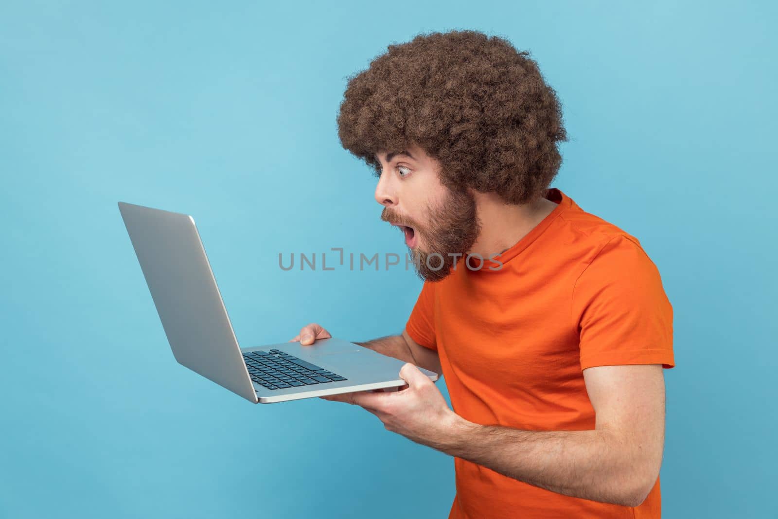 Man with Afro hairstyle in orange T-shirt doing freelance job on laptop, typing email or surfing internet, looking surprised at laptop screen. Indoor studio shot isolated on blue background.