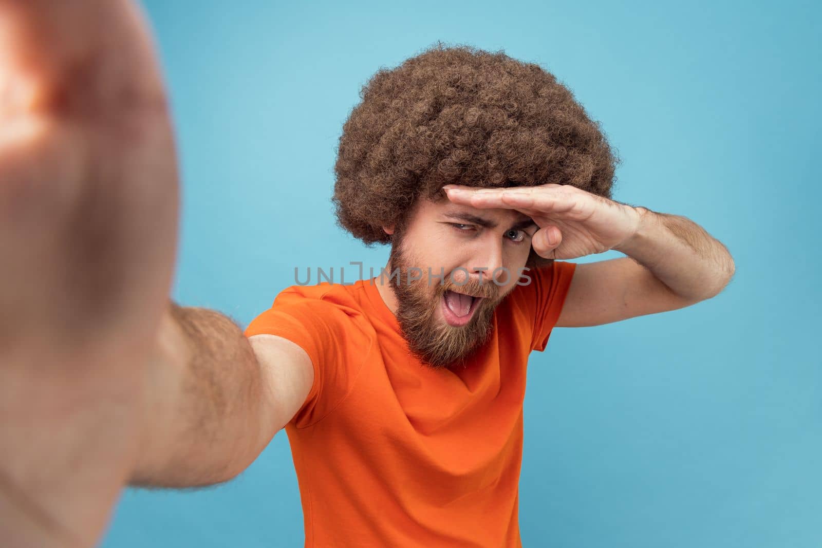 Portrait of positive handsome man with Afro hairstyle wearing orange T-shirt taking selfie for social networks, posing as he is looking far, POV. Indoor studio shot isolated on blue background.