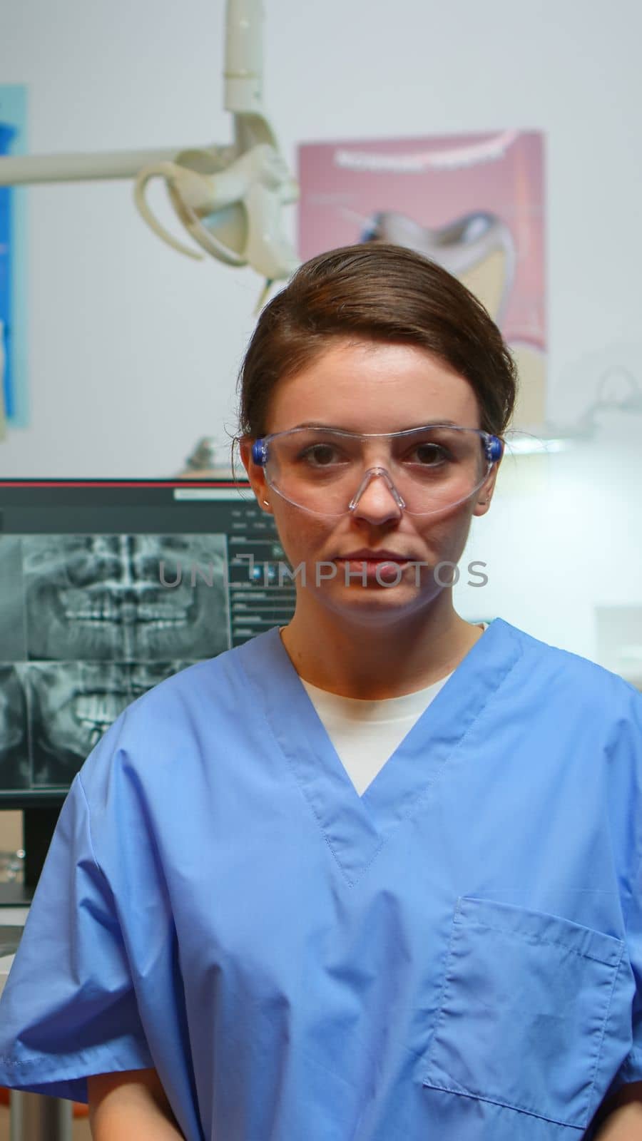 Dental assistant explaining to patients the importance of oral hygiene looking at camera in dental center while doctor is working with patient in background. Stomatologist nurse speaking on webcam