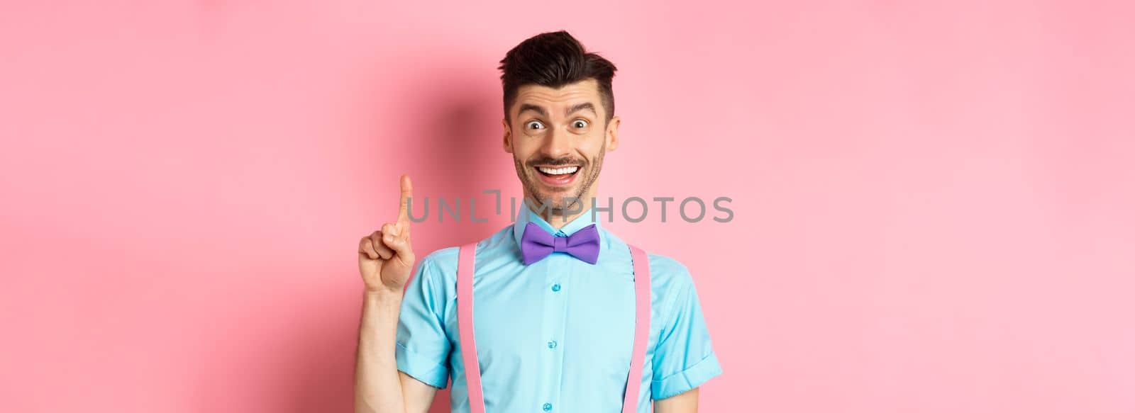 Cheerful caucasian man pitching an idea, raising finger in eureka gesture and smiling, suggesting solution, standing on pink background.