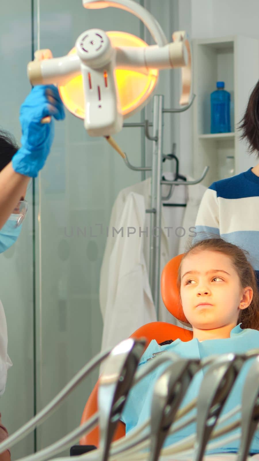 Orthodontist lighting the lamp until examining kid and patient opening mouth. Stomatologist speaking to mother of girl with toothache sitting on stomatological chair while nurse preparing tools.