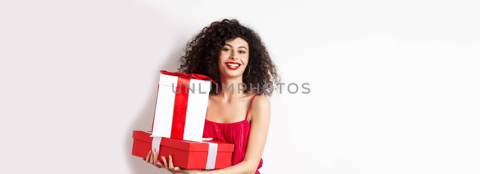 Beautiful birthday girl with curly hair, holding bday gifts and smiling happy, celebrating, standing against white background by Benzoix