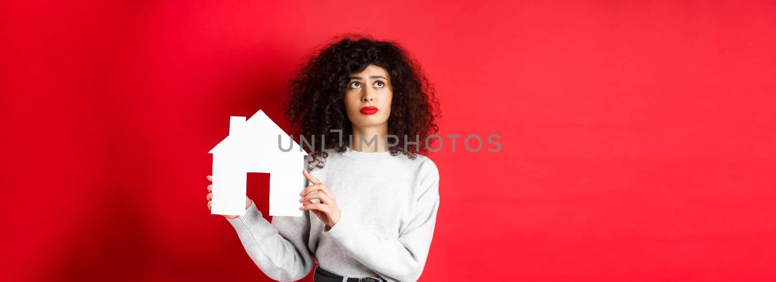 Real estate. Sad woman thinking of buying own house, showing paper home cutout and looking up with uspet face, standing on red background.