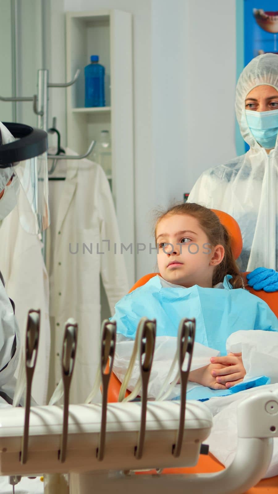 Orthodontist in protection coverall speaking to mother of kid patient with toothache during covid-19 in stomatological office. Concept of new normal dentist visit in coronavirus outbreak