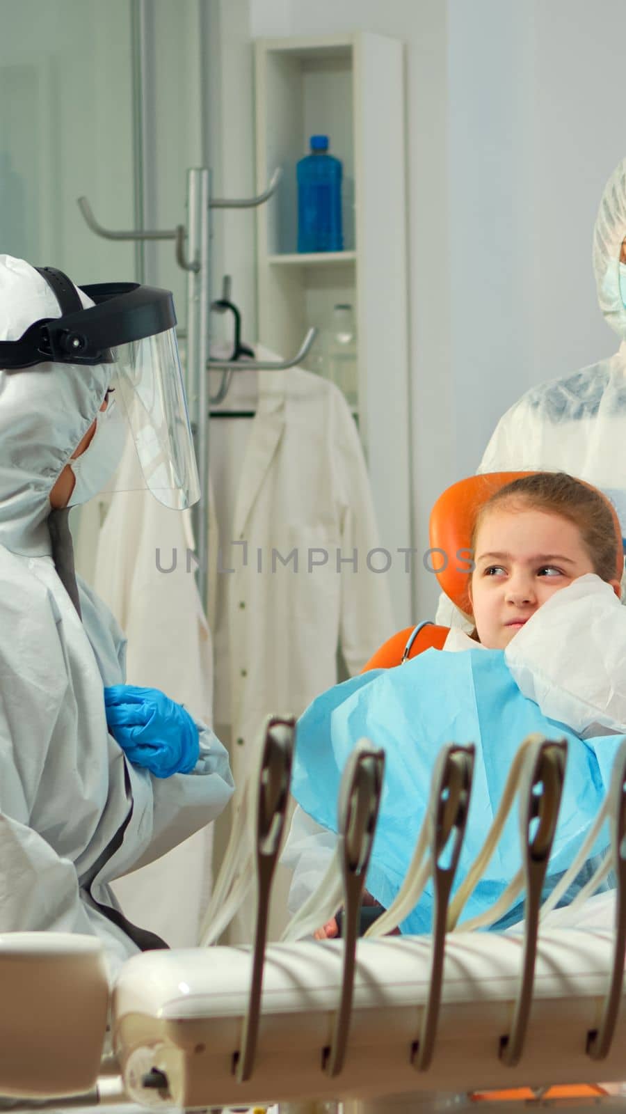 Child in protection suit showing dental problem to orthodontist wearing coverall during global pandemic. Concept of new normal dentist visit in coronavirus outbreak wearing face shield
