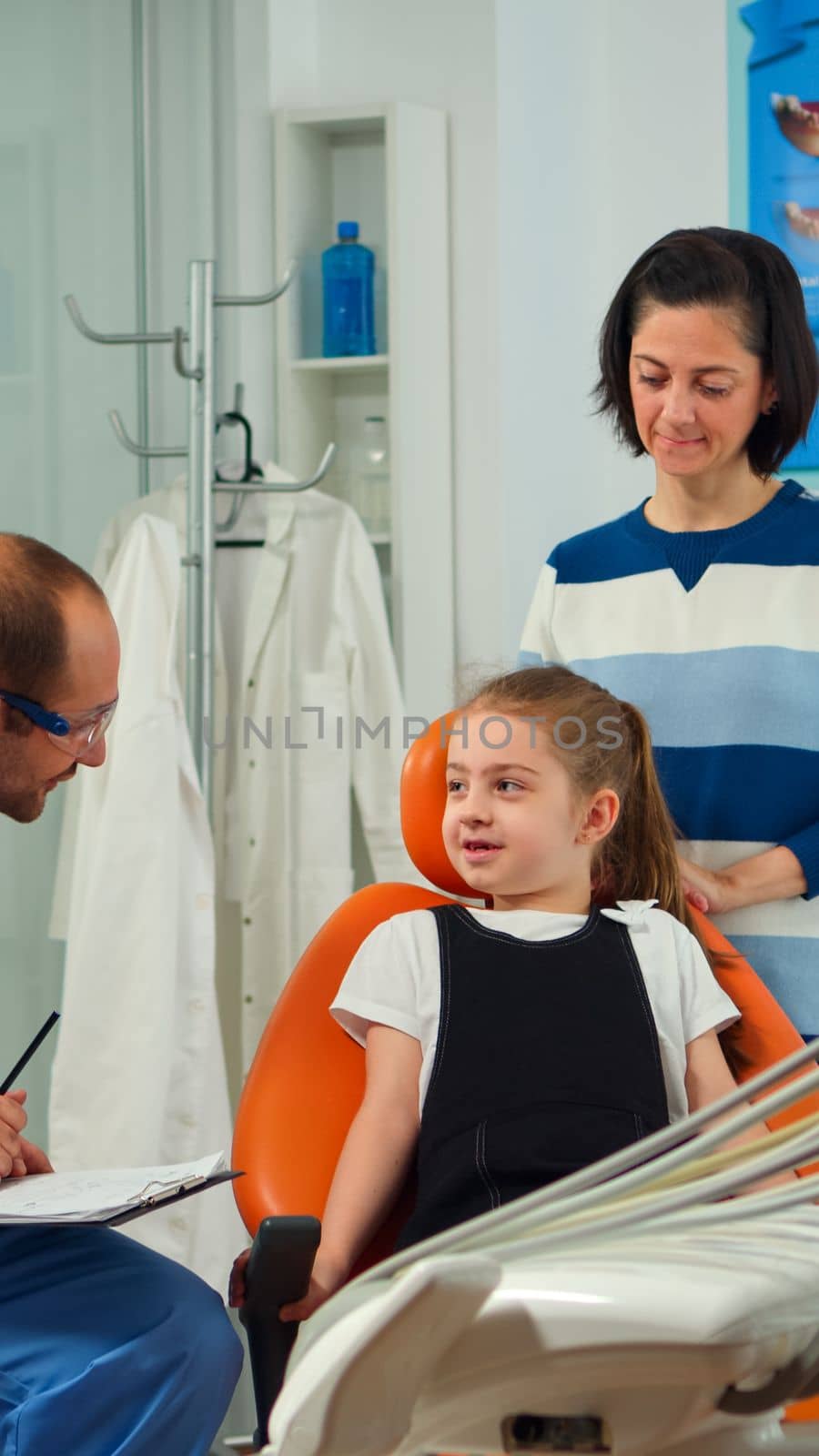 Friendly man dentist assistant interrogating kid patient and taking notes on clipboard waiting for doctor. Mother of little girl explaining dental problem to nurse indicating at toothache problems.