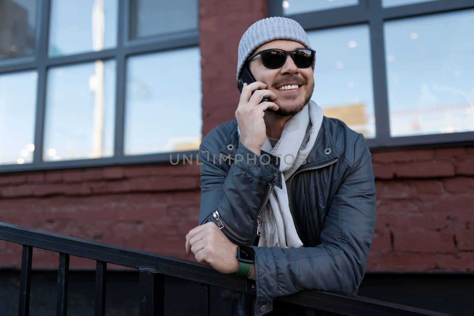 a man in sunglasses and a hat with a scarf speaks with a smile on the phone outside.