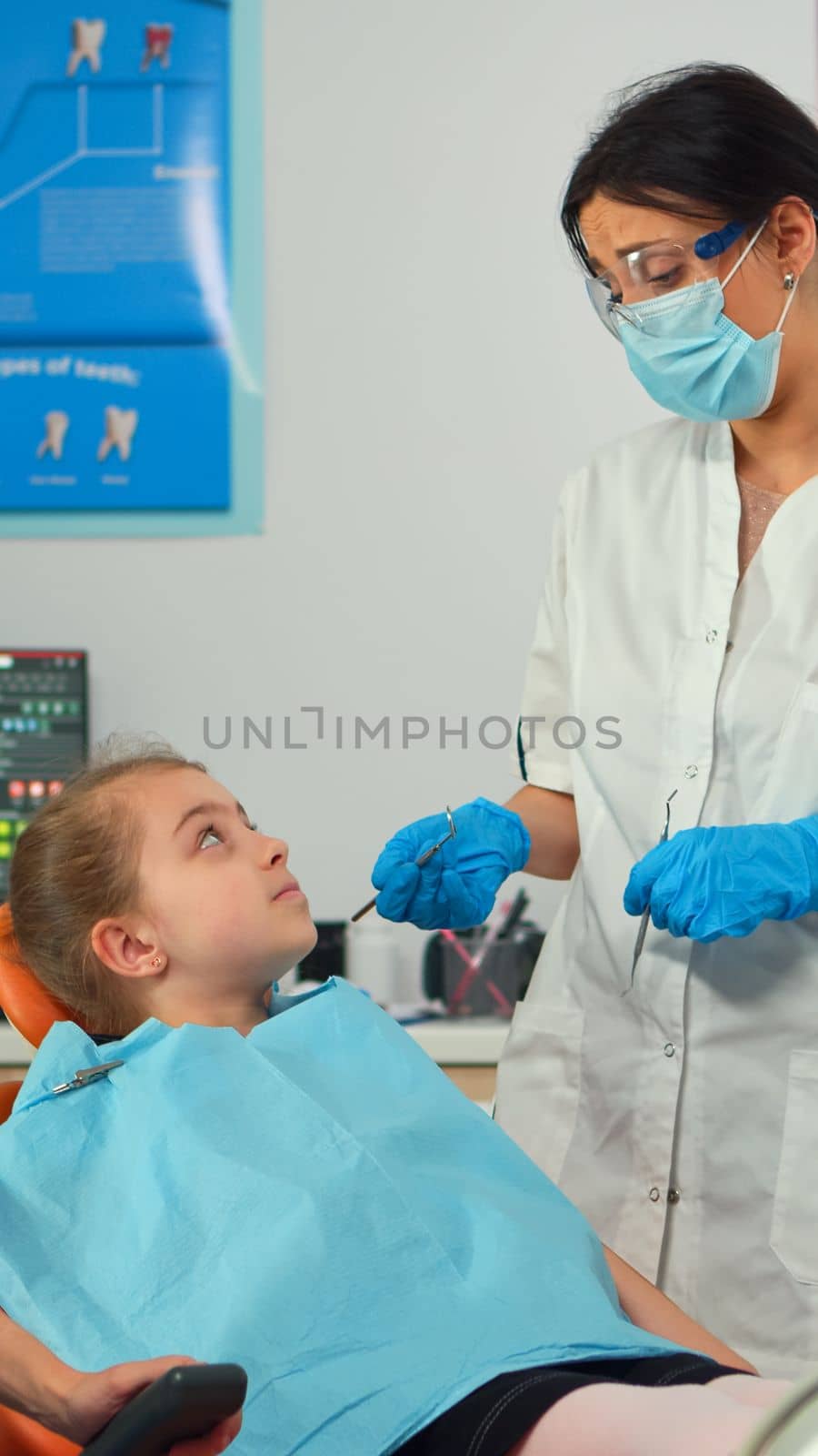 Pediatric stomatologist standing near little girl patient holding stomatological tools talking with mother before intervetion about affected mass. Girl sitting on stomatological chair.