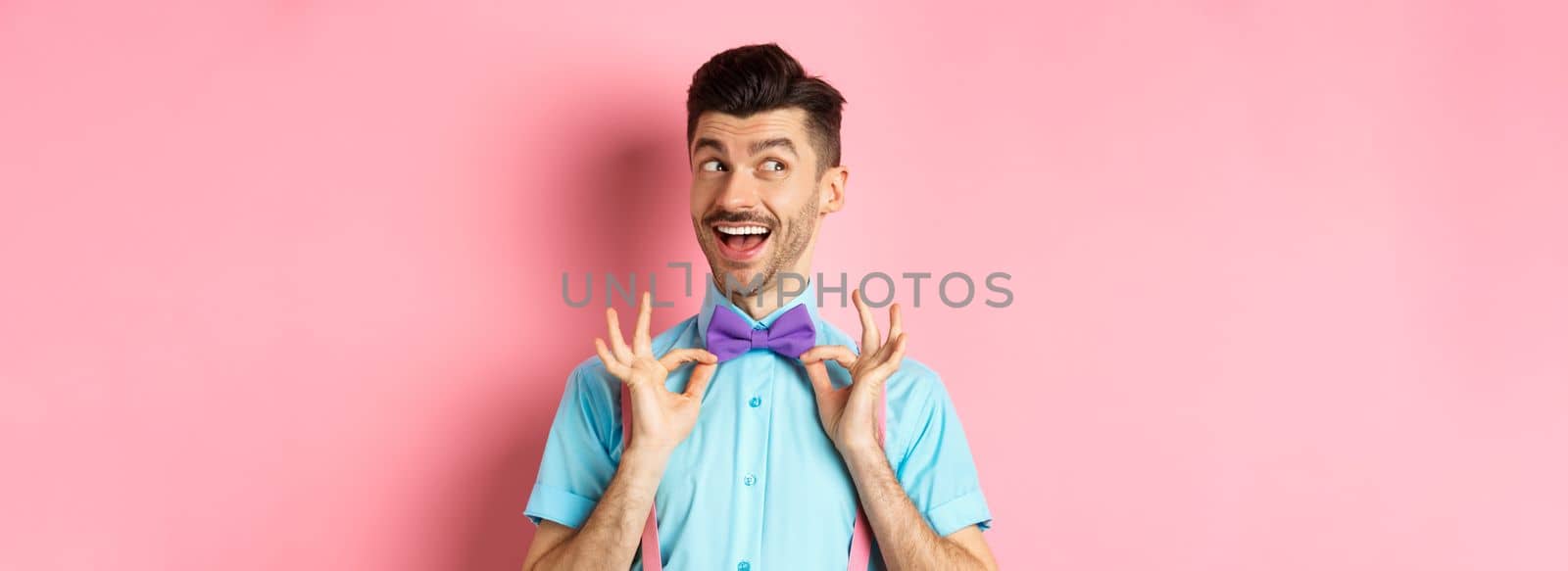 Cheerful and confident young man with moustache, fixing bow-tie on neck and looking left pensive, smiling happy, standing on pink background by Benzoix