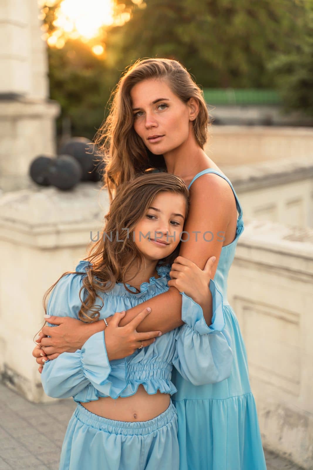 Portrait of mother and daughter in blue dresses with flowing long hair against the backdrop of sunset. The woman hugs and presses the girl to her. by Matiunina