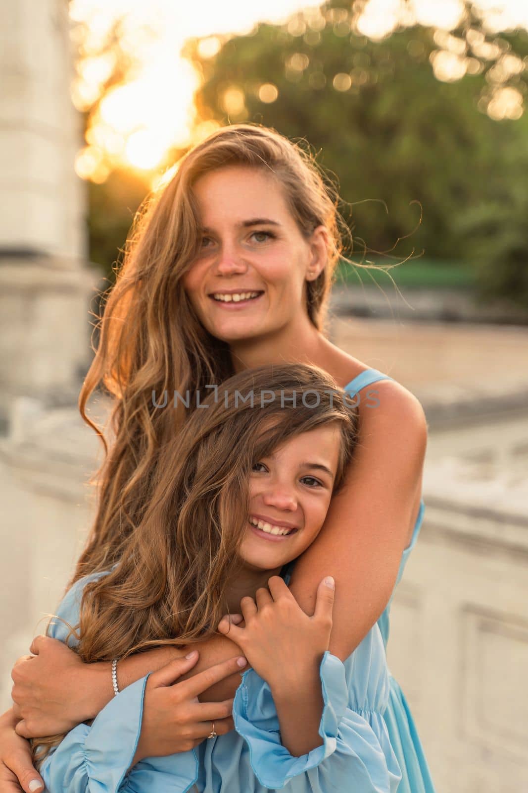 Portrait of mother and daughter in blue dresses with flowing long hair against the backdrop of sunset. The woman hugs and presses the girl to her. They are looking at the camera