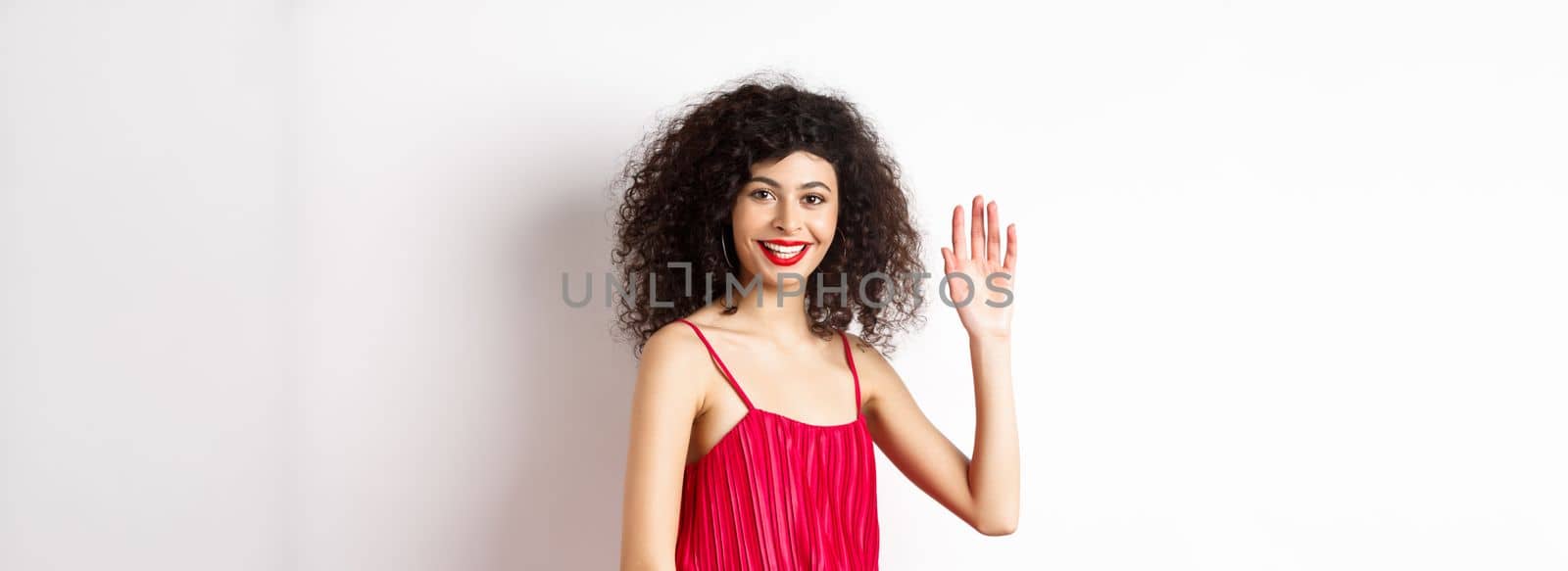 Beauty and fashion. Friendly elegant woman with curly hair, red dress, waving hand and saying hello, smiling to greet you, standing on white background.