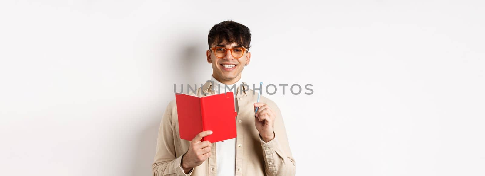 Happy person in glasses writing in journal, holding pen and diary, smiling at camera, plan a schedule, standing with planner on white background.