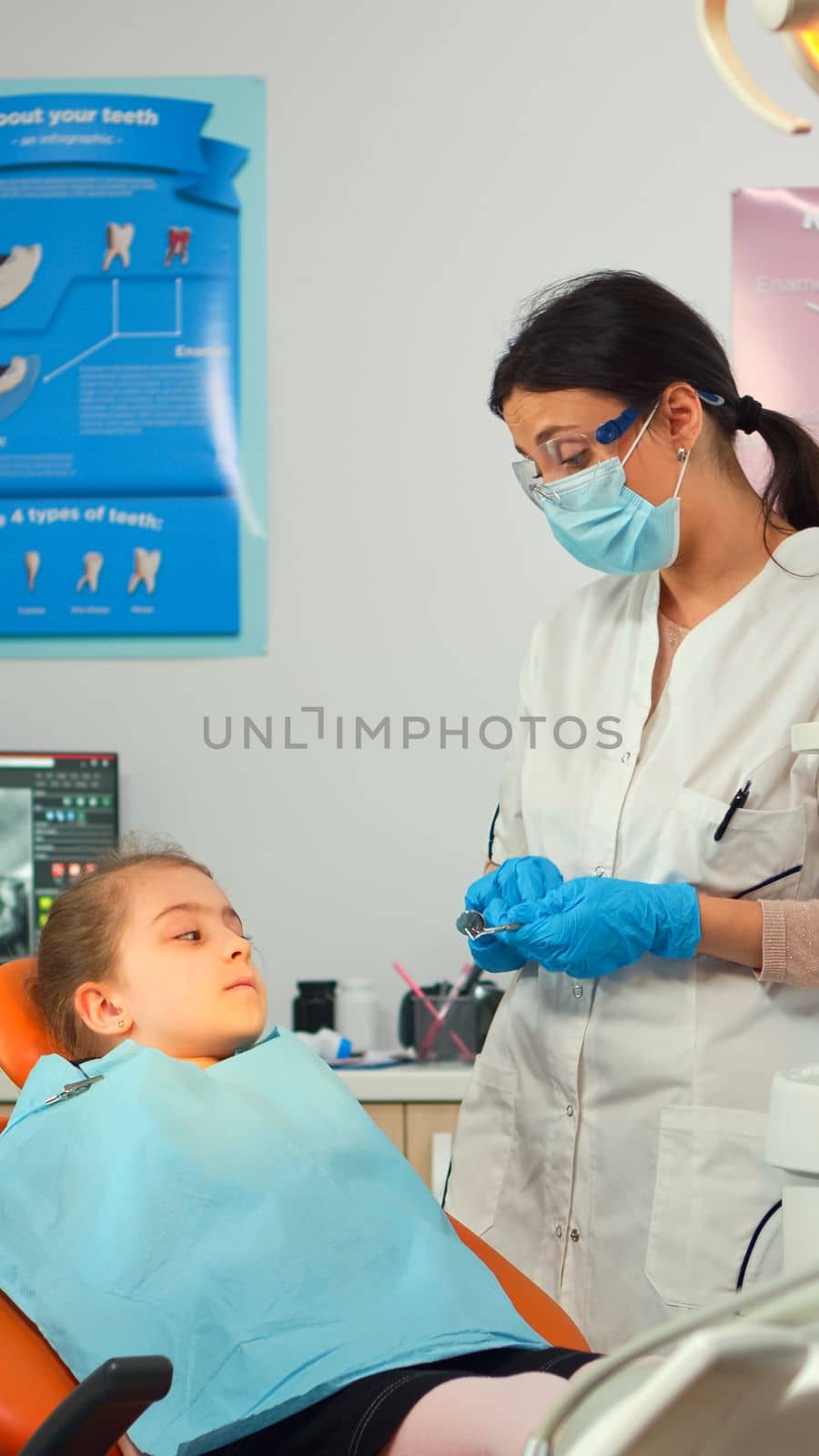 Pediatric dentist with mask checking dental health of a little girl sitting in stomatological chair, doctor using sterilized dental instruments, working with man nurse in modern stomatological unit.