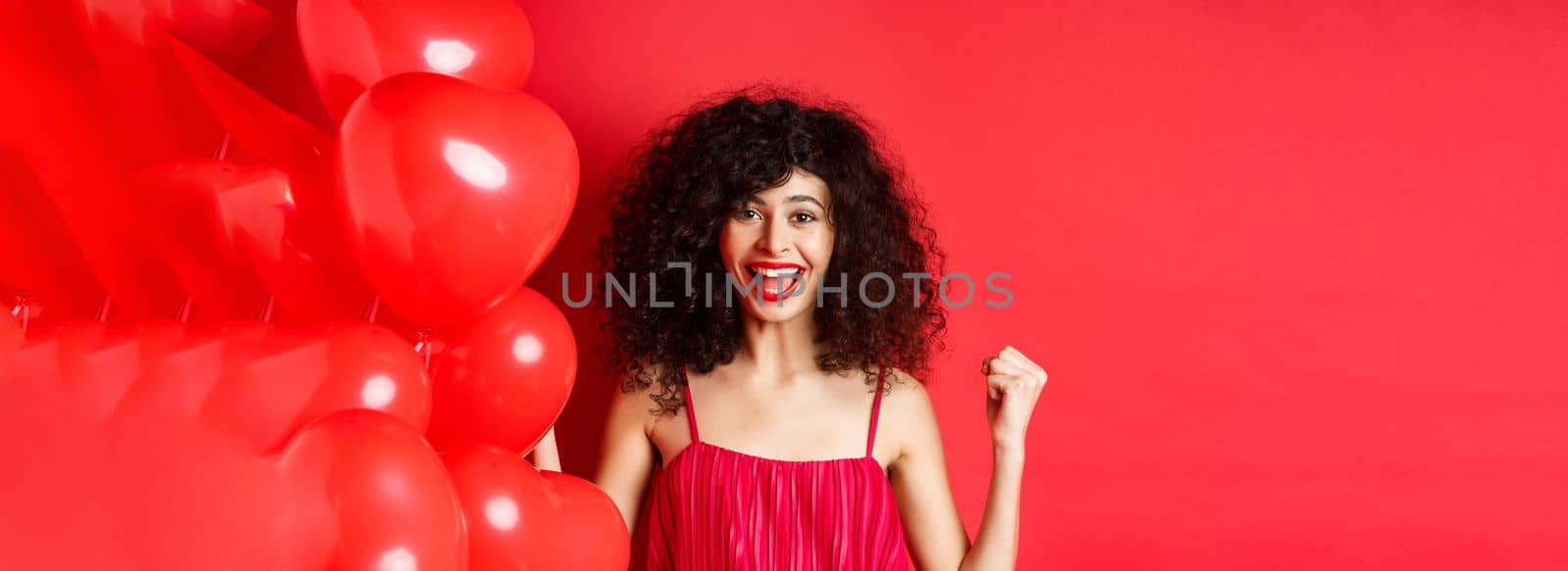 Excited pretty woman winning, say yes and smiling, celebrating success, making fist pump motivated, standing near heart balloons and red background.