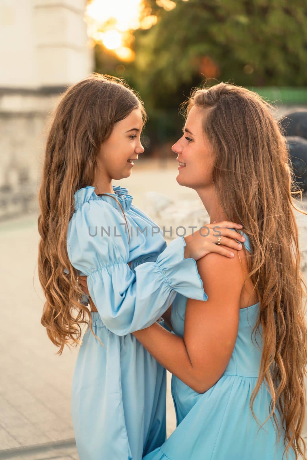 Portrait of a mother and daughter in blue dresses with flowing long hair against the backdrop of a sunset and a white building. They look at each other. Family stories on the weekend.