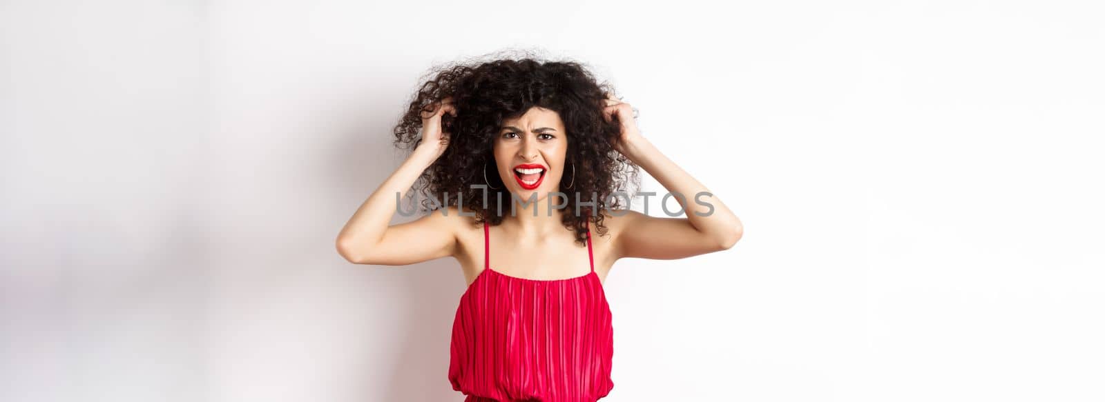 Frustrated curly woman in red dress, frowning and screaming angry, pull out hair and shout at camera, standing on white background.