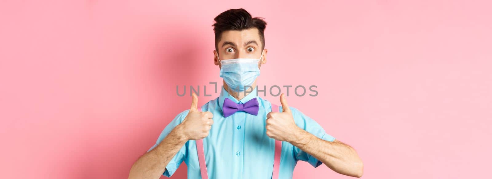 Coronavirus, healthcare and quarantine concept. Satisfied guy in medical mask showing thumbs up in approval, praising excellent choice, pink background.