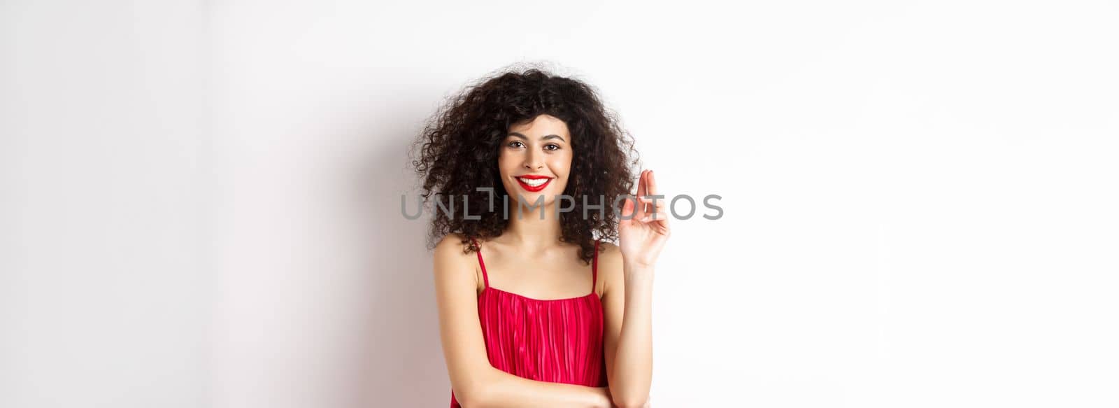 Beauty and fashion. Smiling woman with curly hair and makeup, wearing red dress, waving hand in greeting gesture, saying hello, standing on white background.
