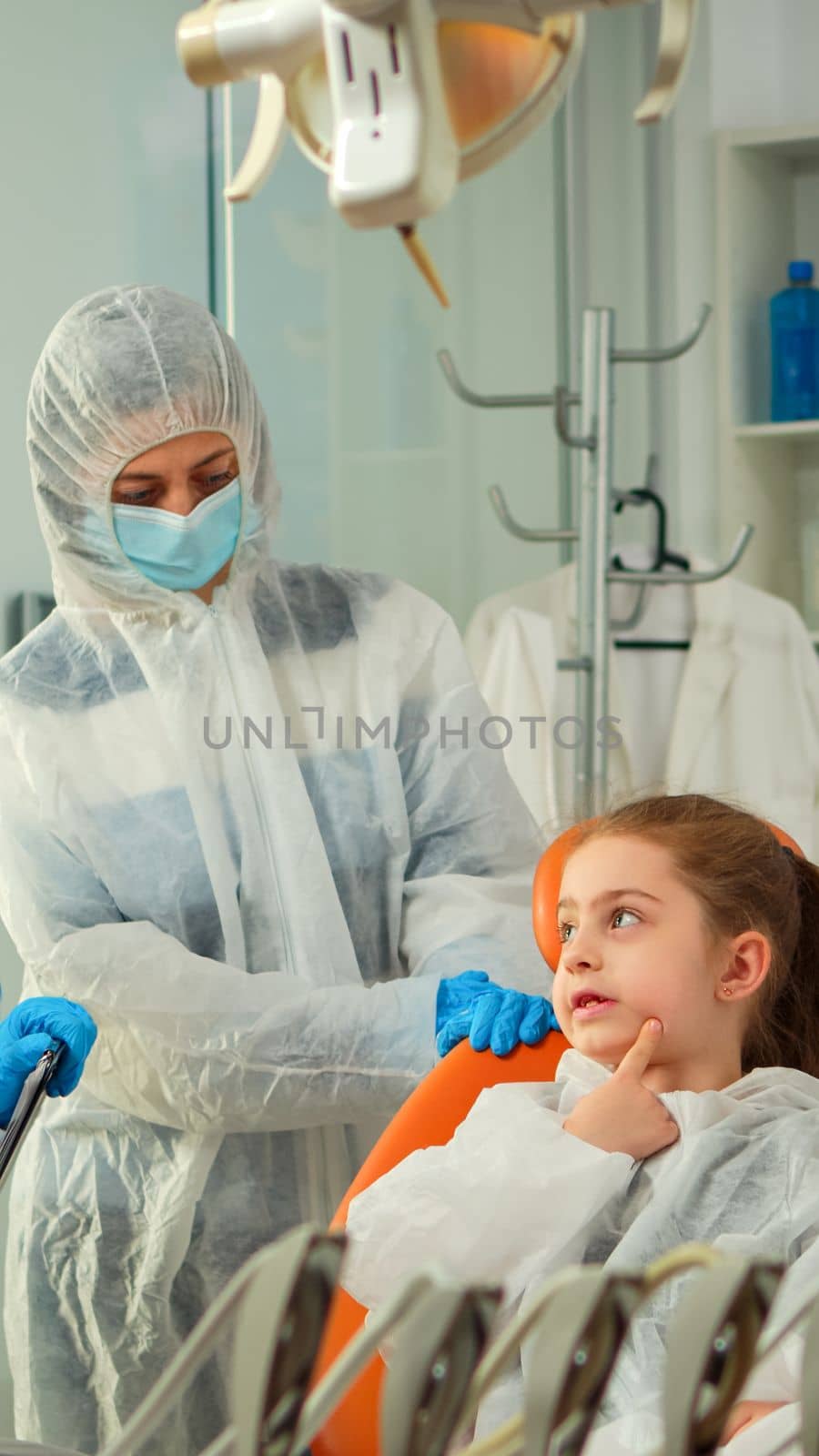Dentist doctor in ppe suit interrogating kid patient and taking notes on clipboard while girl indicating affected mass. Stomatologist and assistent working in new normal dental office wearing coverall
