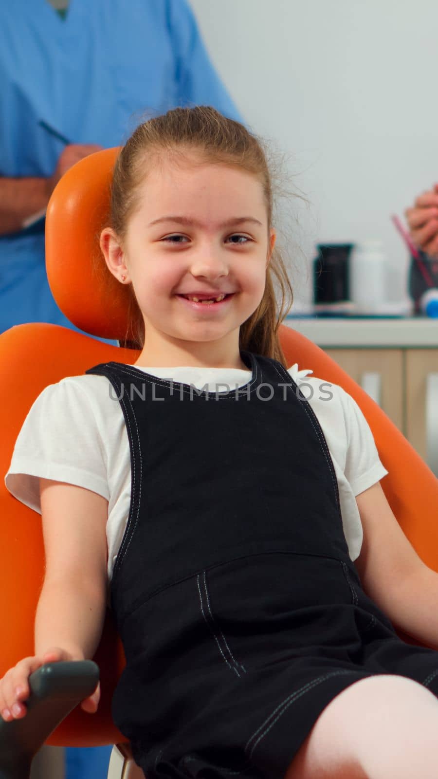 Portrait of little girl patient looking at camera waiting for pediatric stomatologist in dental clinic. Kid lying on stomatologic chair smiling at webcam while nurse and doctor talking with mother.