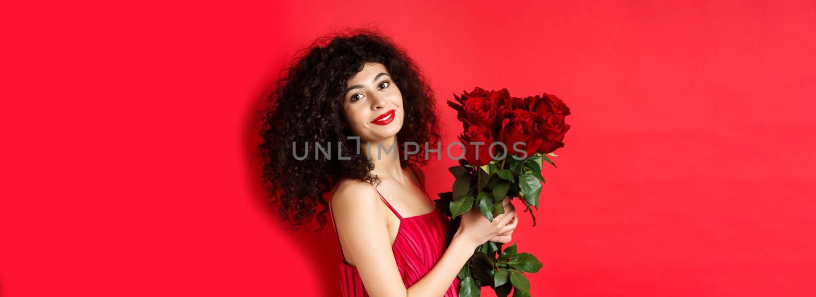 Happy beautiful woman in dress, holding flowers and smiling romantic, standing against red background.